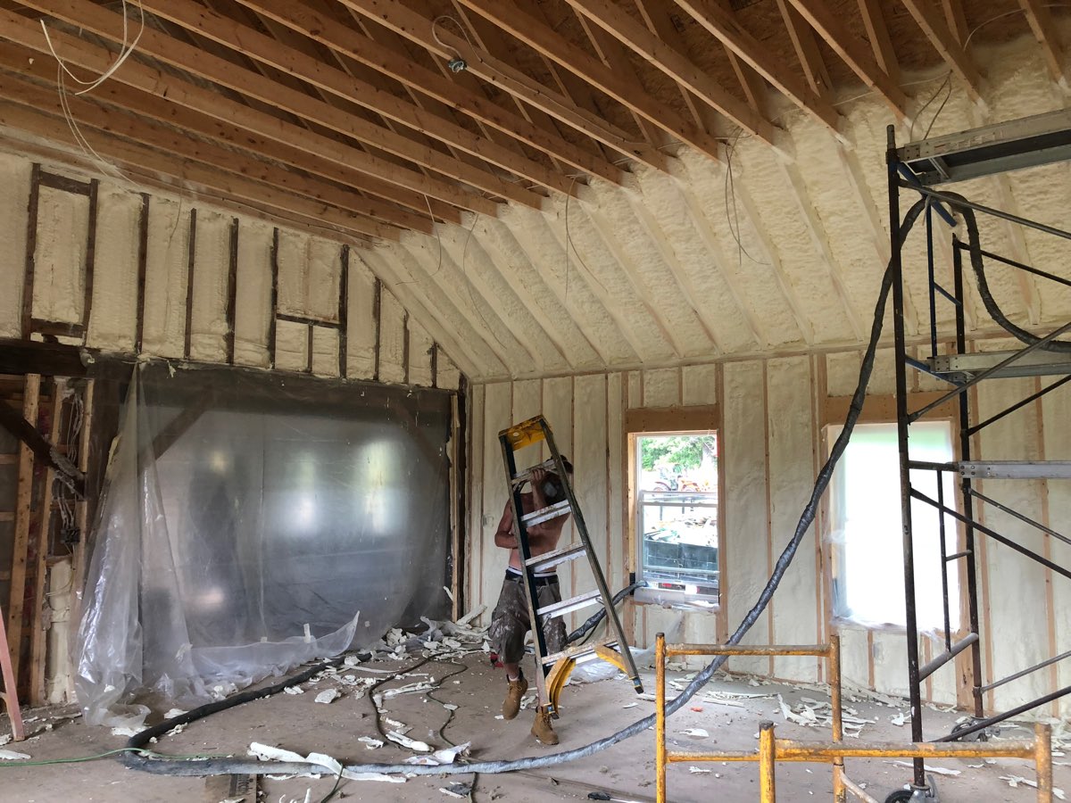 Installer in a respirator moving a stepladder along a vaulted ceiling line freshly sprayed with foam insulation