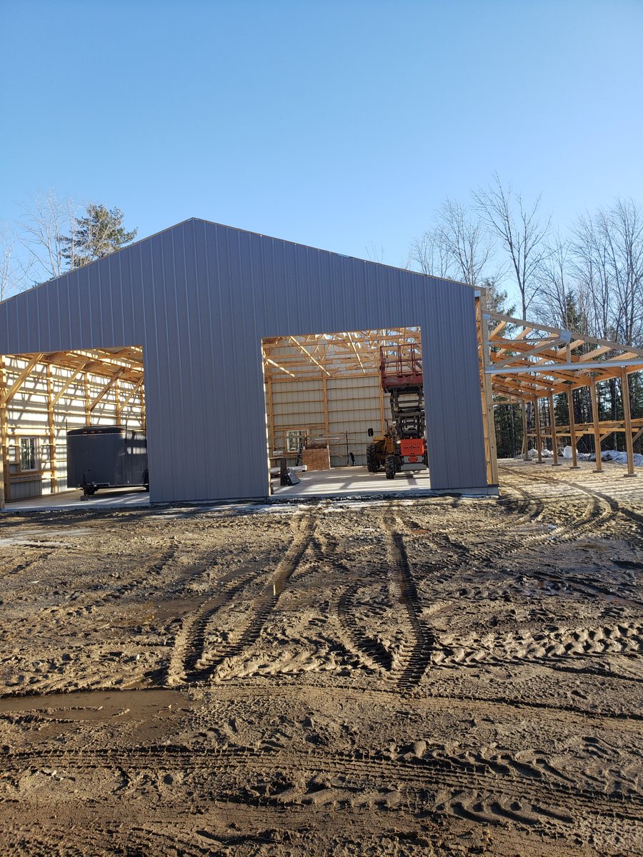 A steel pole barn under construction in Denmark, Maine, with the frame and trusses exposed through the open bay and a scissor lift inside